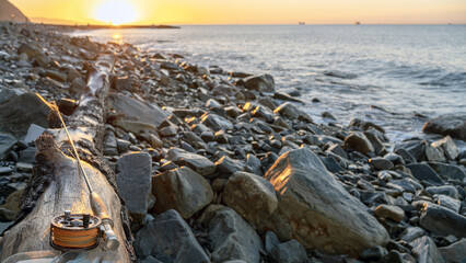 A fly fishing rod and an open fly fishing box lie on the sea rocks at sunset.