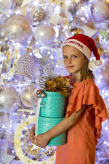 Adorable little girl standing near Christmas tree, wearing Santa hat and holding present gift box. Merry Christmas and happy holidays