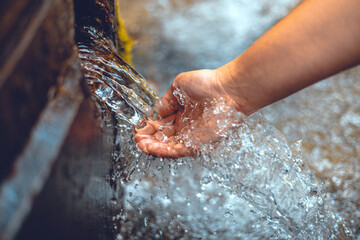 Beautiful photo of a hand under a stream of clear spring water. The woman lowered her hand to touch the stream of clean spring water. The hand touches the cool stream of water. - obrazy, fototapety, plakaty