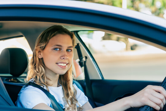the girl is sitting in the car behind the wheel. a young smiling girl sits behind the wheel of a large gray car, a girl with blond wavy hair in a white T-shirt fastened with a seat belt