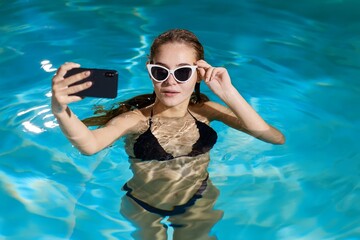 Creative concept. Woman with wet hair and sunglasses takes a photo on her phone in the pool