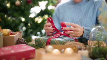 Young asian woman in sweater with happiness to wrapping christmas gift and tie with red ribbon while preparing decorate presents to celebrate for christmas festive holiday and winter seasons at home