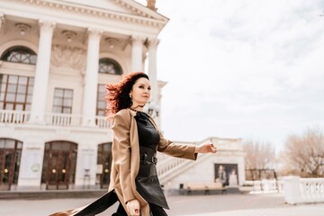 Woman street lifestyle. Image of stylish woman walking through European city on sunny day. Pretty woman with dark flowing hair, dressed in a beige raincoat and black, walks along the building.