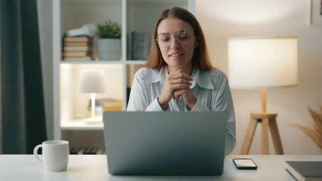 Woman wearing glasses and blue shirt smiling while engaging in live video chat on laptop, discusses work plans with colleagues