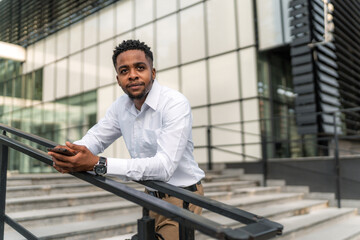 A portrait of a handsome Black businessman leaning against a railing during his break, holding a phone in his hand. 