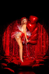 Valentines day. Beautiful Sexy girl model in gift bow outfit joying with air hearts balloons over red studio party background. Slim blond girl. Glamorous style.