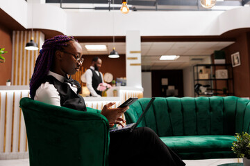 Young African American woman hotel manager sitting at reception lounge area using smartphone and...