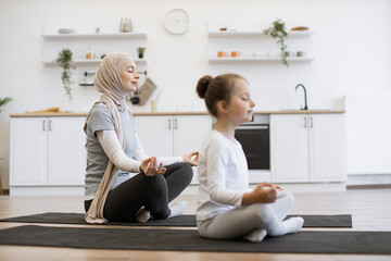 Fototapeta premium Side view focus on peaceful young woman in hijab meditating in seated asana with adorable girl indoors. Calm mother and little daughter working out on yoga mats together on sunny day at home.