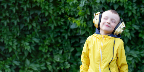 A positive little boy of 6 years old in a yellow jacket and yellow retro earphones listens to music. The boy stands against the background of a wall of plants. Banner with space for text