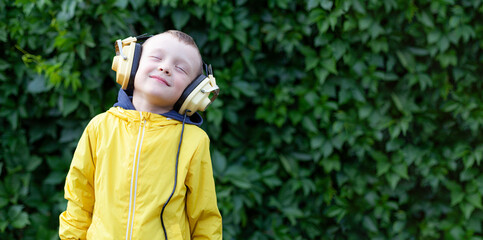 A positive little boy of 6 years old in a yellow jacket and yellow retro earphones listens to music. The boy stands against the background of a wall of plants. Banner with space for text