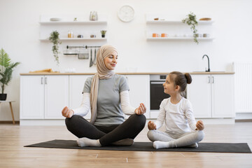 Full length view of relaxed muslim female sitting in yoga pose with little child on rubber mat in modern apartment. Fit mother exercising with fingers in gyan mudra with daughter looking at each other