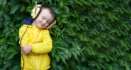 A positive little boy of 6 years old in a yellow jacket and yellow retro earphones listens to music. The boy stands against the background of a wall of plants. Banner with space for text