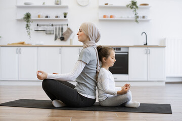 Energetic little girl sitting back to back with muslim lady during her achieving cross-legged position. Happy mother promoting daughter's sense of calmness by meditating together at home.