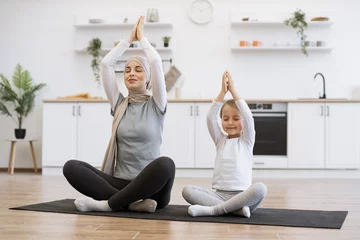 Foto auf Leinwand Beautiful adult muslim woman and her cute daughter in activewear taking up lotus posture on mats in meditation room. Mother and little girl improving balance and flexibility while exercising at home. © sofiko14