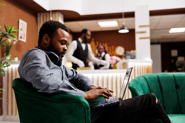 Focused African American man freelancer typing on laptop keyboard using hotel wifi, sitting in...