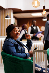 Happy smiling African American woman arriving at resort, sitting in hotel lobby with smartphone and...