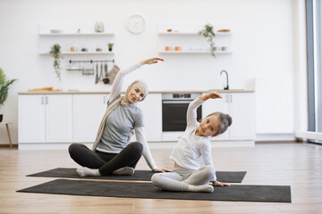 Slim muslim lady on yoga mat stretching arms while training with small female kid. Young parent and daughter in sportswear spend time happily during workout.