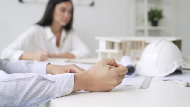 Professional architect engineer drawing blueprint and working together with young beautiful caucasian coworker on meeting table with safety helmet, house model, blueprint scatter around. Immaculate.