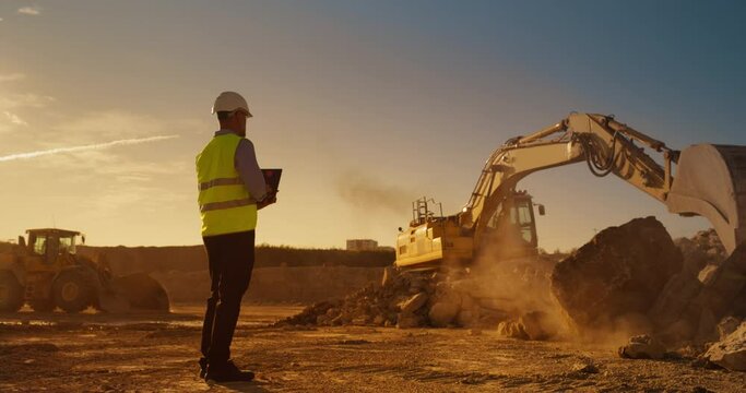 Caucasian Male Inspector Using Laptop Computer On Construction Site Of New Real Estate Project. Man Observing Industrial Machinery At Work, Laying Foundation For Apartment Building. Sunny Evening