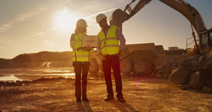 Hispanic Female Urban Planner Using Laptop And Talking To Caucasian Male Real Estate Developer On Construction Site. Colleagues Discussing Building Project, Excavator Working On Background. Warm Edit