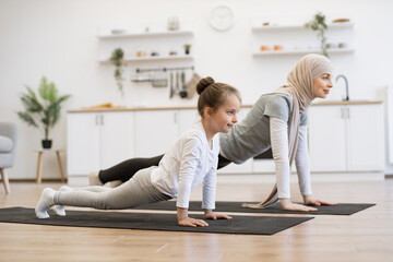 Attractive muslim mother in hijab practicing plank position with baby daughter in kitchen after wake up in morning. Feeling so comfortable and relax. Yoga of woman with her baby girl for healthcare. - obrazy, fototapety, plakaty