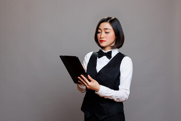 Confident asian waitress using digital tablet and checking orders online while posing in studio. Young woman restaurant receptionist wearing professional uniform holding portable gadget