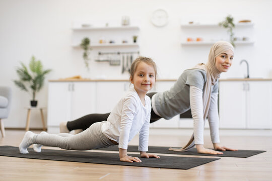 Attractive muslim mother in hijab practicing plank position with baby daughter in kitchen after wake up in morning. Feeling so comfortable and relax. Yoga of woman with her baby girl for healthcare.