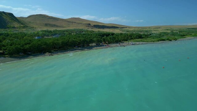 Aerial view of a large blue azure sea lake in clear sunny weather. People are swimming on the beach and pine trees are growing, boats are standing. Drone video, flying in an arc. Lake Sevan, Armenia.