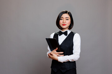 Confident young asian woman waitress wearing uniform and bow tie standing with digital tablet...