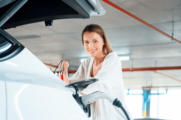 Young woman travel with EV electric car to shopping center parking lot charging in downtown city...