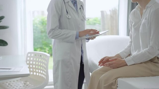 Doctor and patient are in the clinic. Pediatrician is standing and using a tablet PS for making medical notes while a young woman is sitting on a sofa and talking. Medicine, healthcare concepts