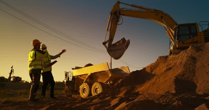 Caucasian Real Estate Investor With Tablet Computer And Project Manager Talking On Construction Site Of Apartment Building. Colleagues Wearing Hard Hats And Watching Heavy Machinery Working. Sunny Day
