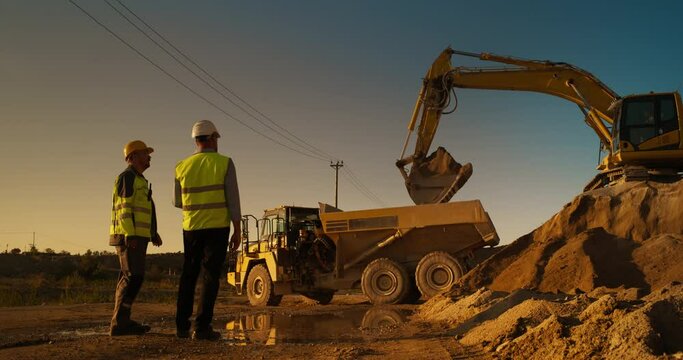 Caucasian Male Real Estate Investor And Project Manager Talking On Construction Site Of Apartment Block. Colleagues Discussing Building Progress. Excavator Loading Sand In Industrial Truck On Warm Day