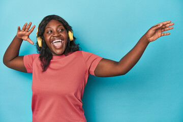 Joyful curvy woman dancing with headphones on blue backdrop