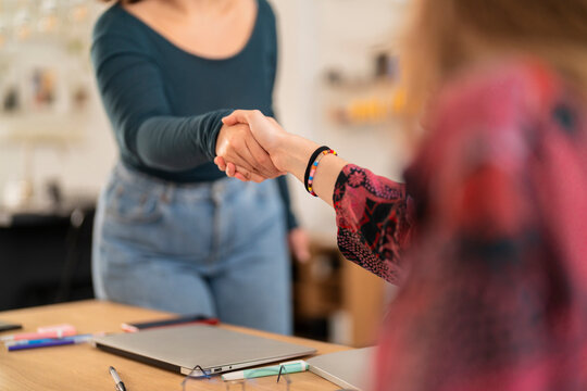 Crop women shaking hands and greeting each others in modern workspace at office table against blurred background of smiling female colleague