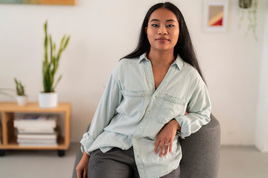 Young Asian female wearing casual clothes looking at camera while leaning chair in light office against blurred background