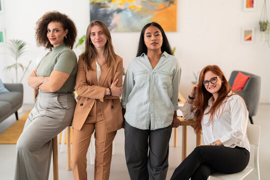 Smiling multiethnic females colleagues in casual outfit looking at camera while leaning on table with hand on shoulder in modern office