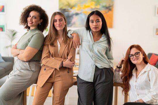 Smiling multiethnic females colleagues in casual outfit looking at camera while leaning on table with hand on shoulder in modern office