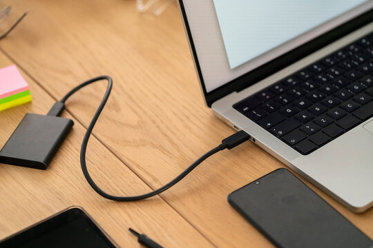 High angle of crop anonymous female sitting at wooden table and plugging external HDD in laptop with white screen and stationery in office