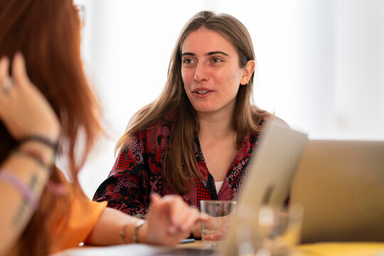 Serious female in casual clothes with laptop at wooden table and looking away while having pen in hand while sitting against blurred background at office