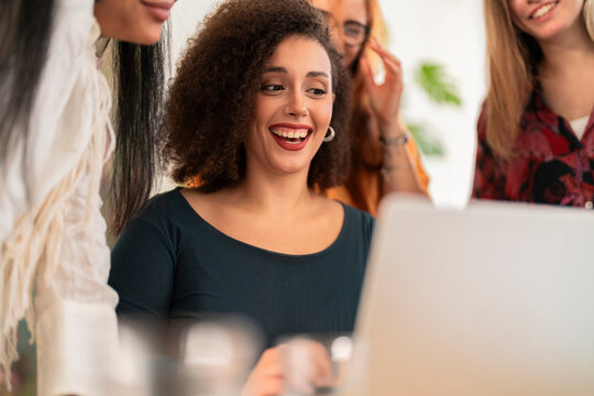Positive female worker with crop multiracial colleagues in casual clothes gathered at table with laptop and smiling while looking at screen and working together in office