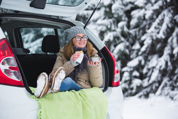 Woman in woolen hat sits in the trunk of the car and holds a cup of hot tea in her hands