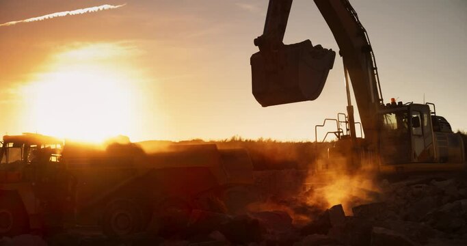 Cinematic Golden Hour Shot Of Construction Site: Excavator Loading Big Rocks Into Industrial Truck. Two Workers Discussing Progress Of Building New Real Estate Project. Real Estate Development Concept