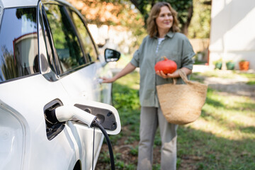 Woman with shopping bag and pumpkins next to a charging electric car in the yard of a country house.