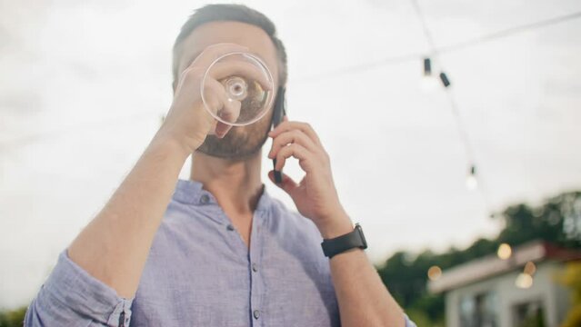 Portrait of carefree young man standing and talking by phone outdoors . Handsome man drinking alcohol from wineglass celebrating holiday at street party. Summertime free time and holiday concept.