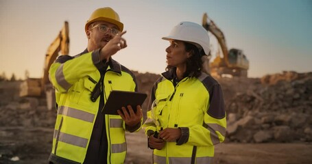Caucasian Male Civil Engineer Talking To Hispanic Female Inspector And Using Tablet On Construction Site of New Apartment Complex. Real Estate Developers Discussing Business, Excavators Working.