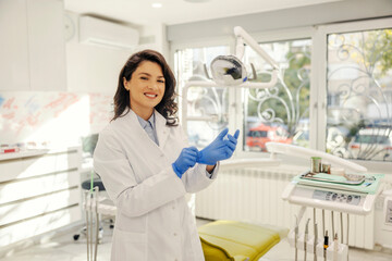A happy dentist is putting on gloves while standing at dentist office.