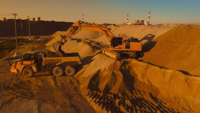 Aerial Drone Shot Of Construction Site On Sunny Day: Industrial Excavator Loading Sand Into A Truck. The Process Of Building New Apartment Block. Workers Operating Heavy Machinery Concept.