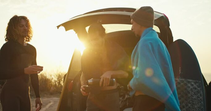 Three guy friends pour tea from a thermos into special cups and warm up after surfing and swimming in cold water at sunrise. Guys drinking tea near their black car at Sunrise