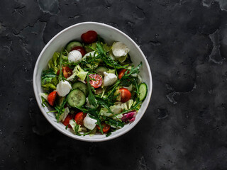 Arugula, cherry tomatoes, cucumbers, mozzarella cheese salad with lemon-mustard dressing on a dark background, top view